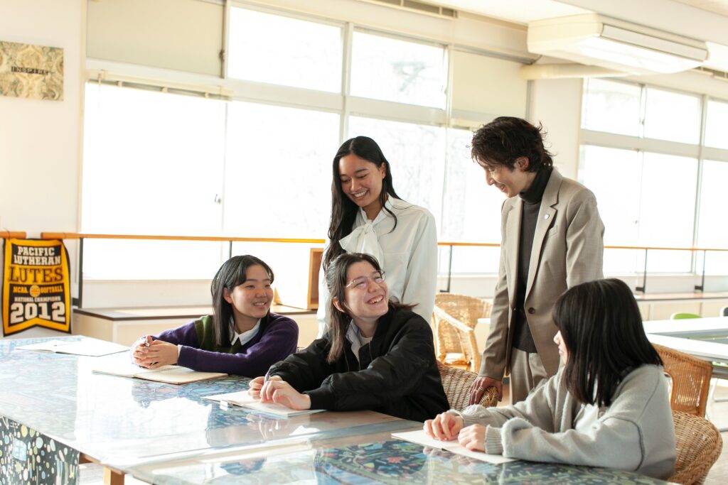 AIE国際高等学校神戸キャンパスの明るい教室で、先生と多様な生徒たちが笑顔で交流しながら、少人数制の手厚いサポートを受けて学ぶ授業風景。