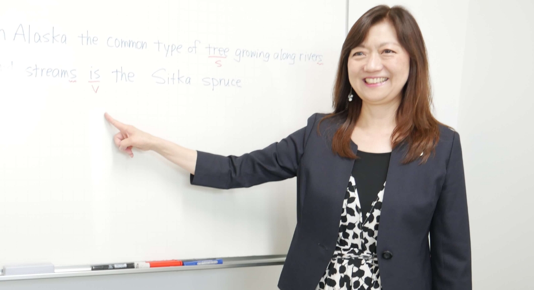AIE国際高等学校 神戸キャンパスで、ホワイトボードの英文を指差しながら笑顔で実践的な英語の授業を行う女性講師。